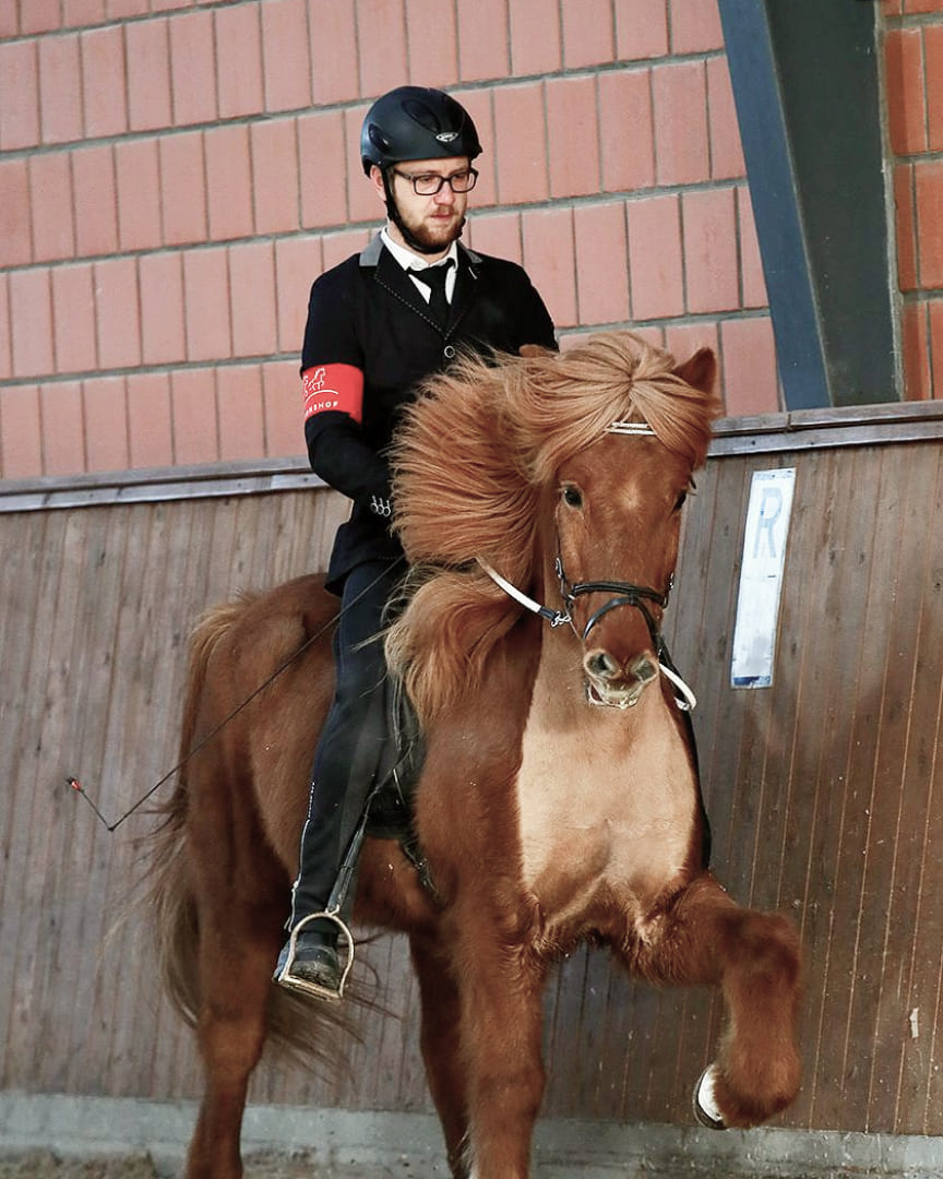 Fabian-Schrader-Ueber-mich-3 Ein Reiter in Turnierkleidung reitet ein fuchsfarbenes Islandpferd mit wehendem Langhaar in einer Reithalle.