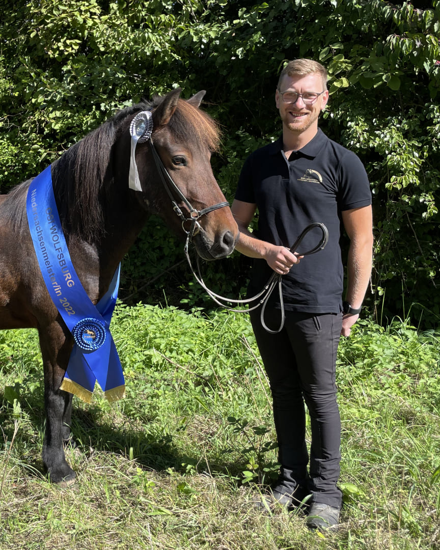 Fabian-Schrader-Islandpferde- new2025-2 Ein Mann in schwarzem Poloshirt steht lächelnd neben einem prämierten Islandpferd mit blauer Siegerschärpe und silberner Schleife.